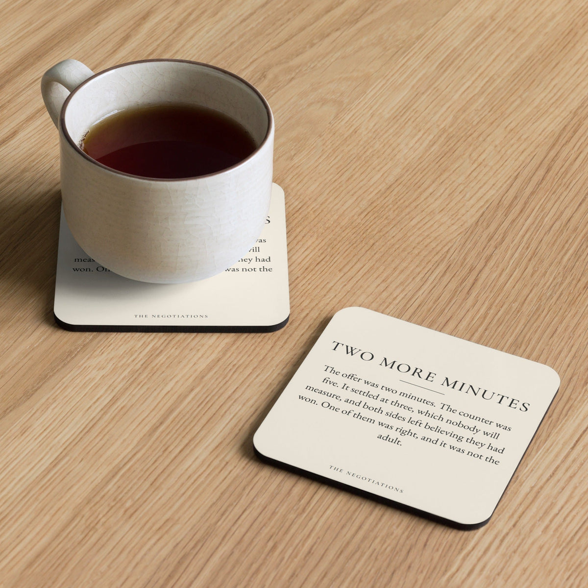 The Two More Minutes coaster from The Negotiations on a wooden table with a cup of tea